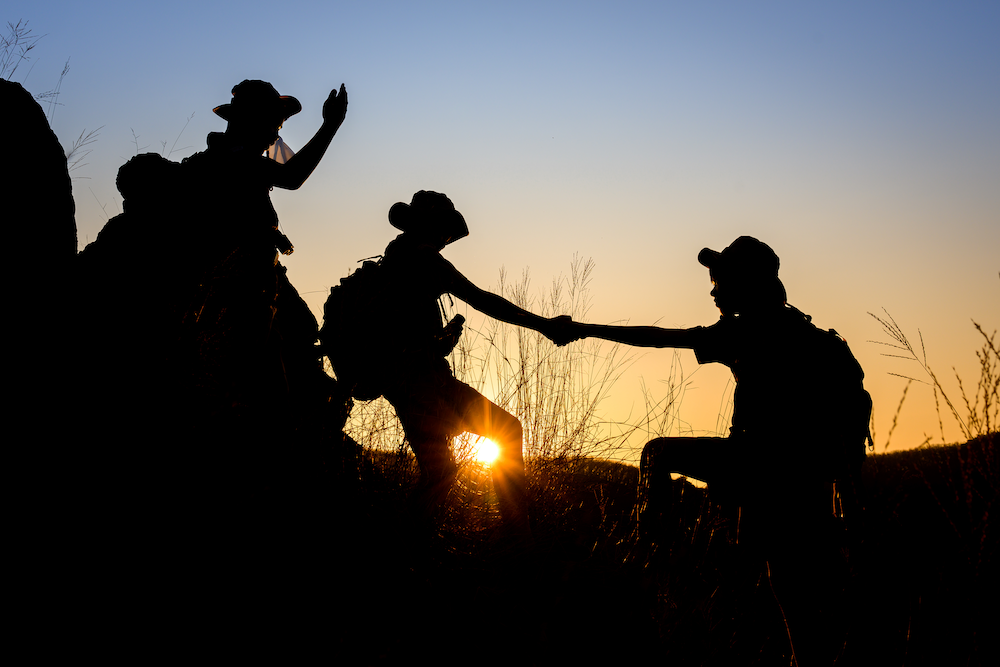 silhouette of Scout students showing help to each other Hold hands and pull each other up from the cliff to explore the beautiful forest in the evening as the sun sets.